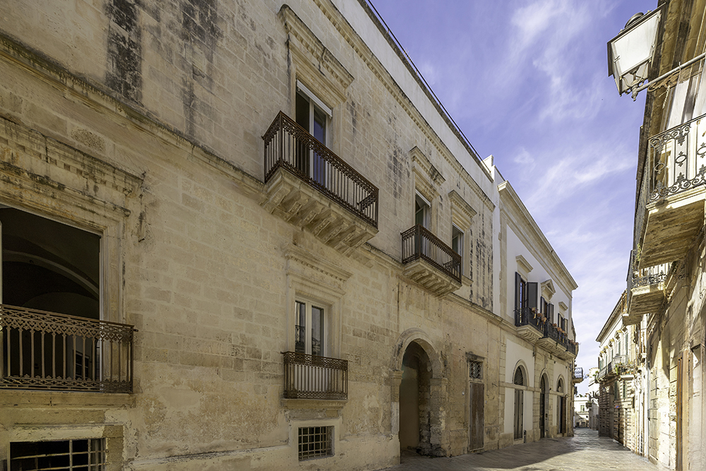 Low-angle view of a historic beige stone building with ornate iron balconies along a narrow, sunlit Italian street. [AI-generated text]