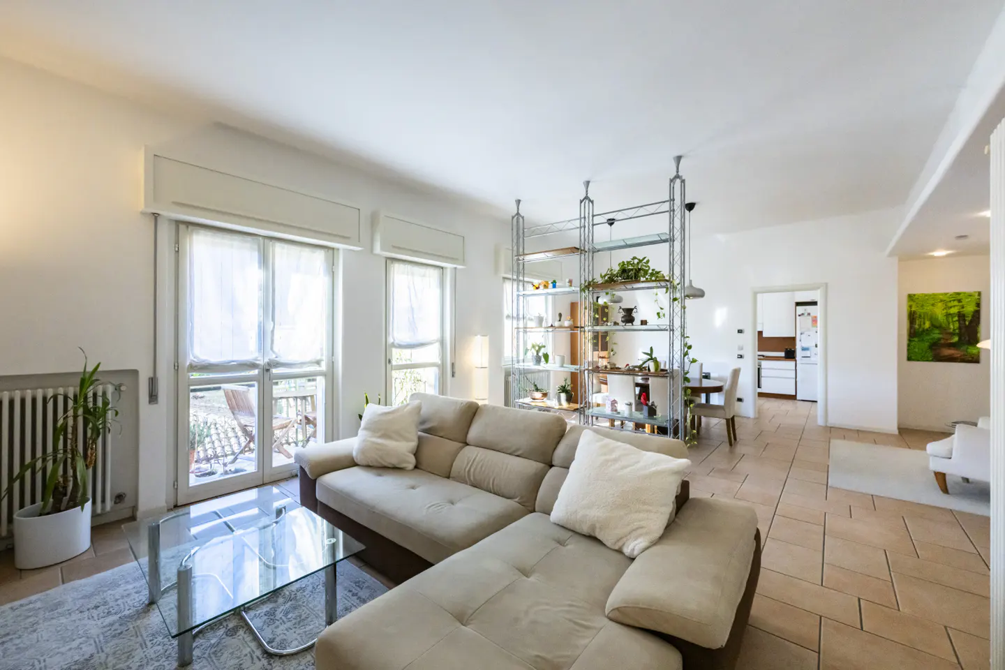 Bright living room with beige sectional sofa, glass coffee table, and metal shelving unit with plants. Tiled floor and white walls.