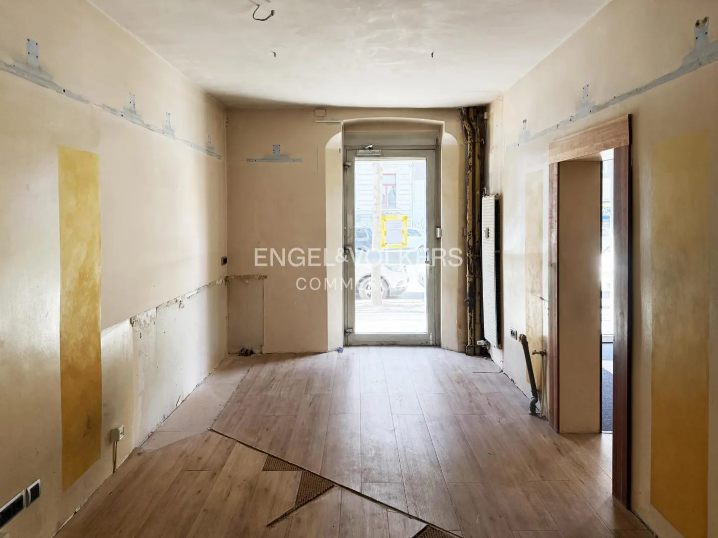 Unfinished commercial space with light wood floors, beige walls, and a glass door leading to the street. Engel & Volkers logo visible.