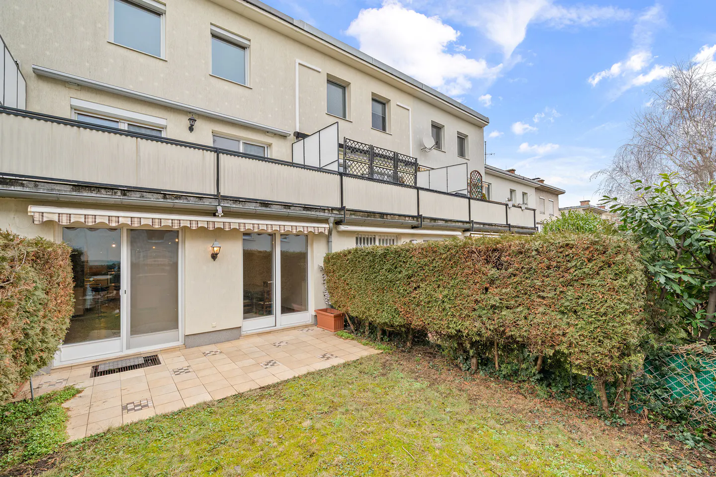 Backyard view of a multi-story beige building with sliding glass doors, a patio, and a green lawn.