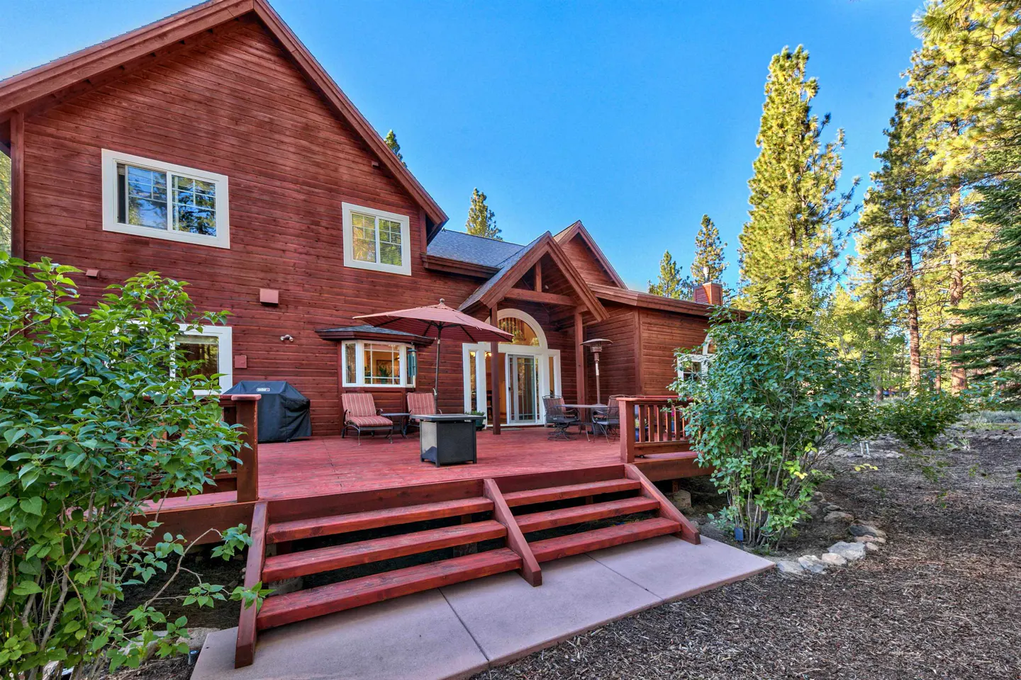 Red wooden house with a deck, umbrella, grill, and outdoor seating. Trees and blue sky in the background.