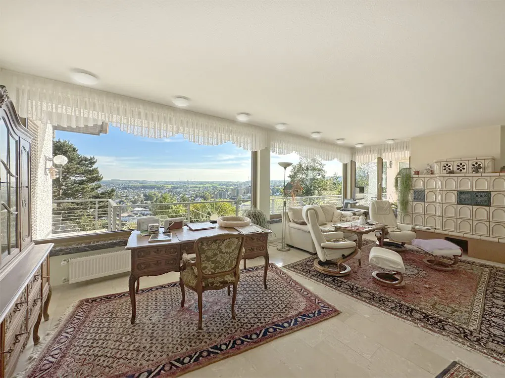 Bright living room with a desk, chairs, and a white tiled fireplace. Large windows offer a scenic view of the city and landscape.