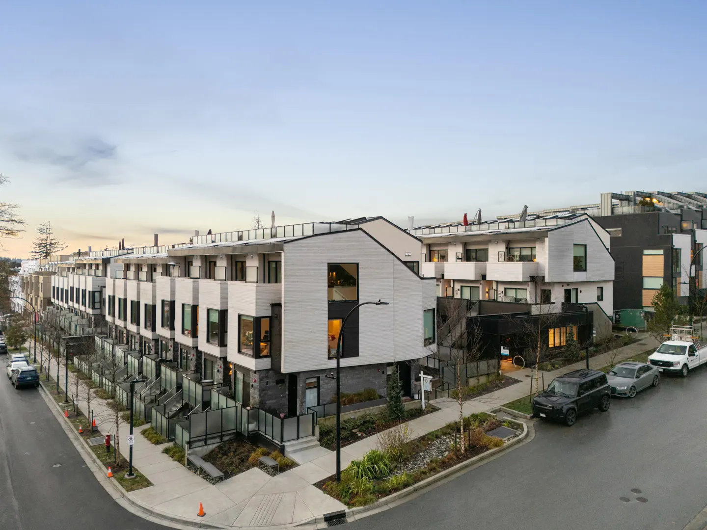 Modern townhouses line a street, featuring white and gray exteriors, black-framed windows, and rooftop terraces under a pale sky.