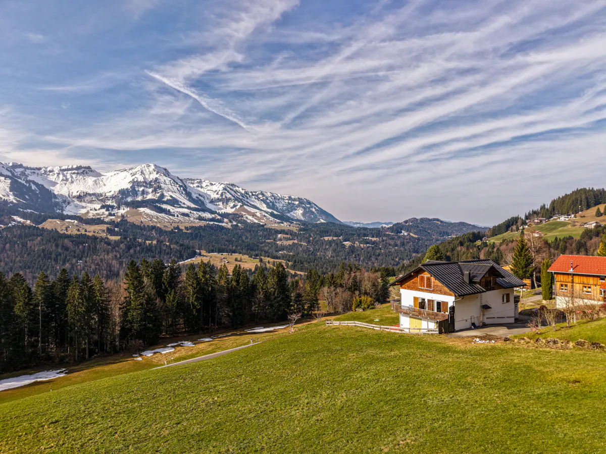 Scenic view of a white house with a black roof on a green hillside, with snow-capped mountains and a blue sky in the background.