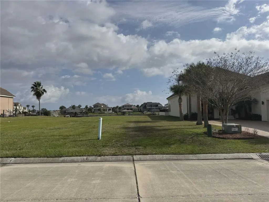 Vacant grassy lot with waterfront views. Houses line the water in the background under a partly cloudy sky.