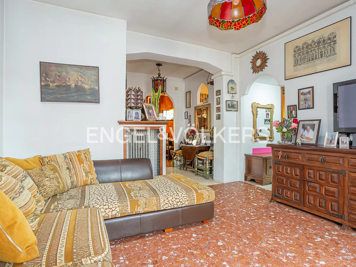 A living room with a brown leather couch, patterned cushions, and a red-tiled floor. Archways lead to other rooms, and artwork adorns the white walls.