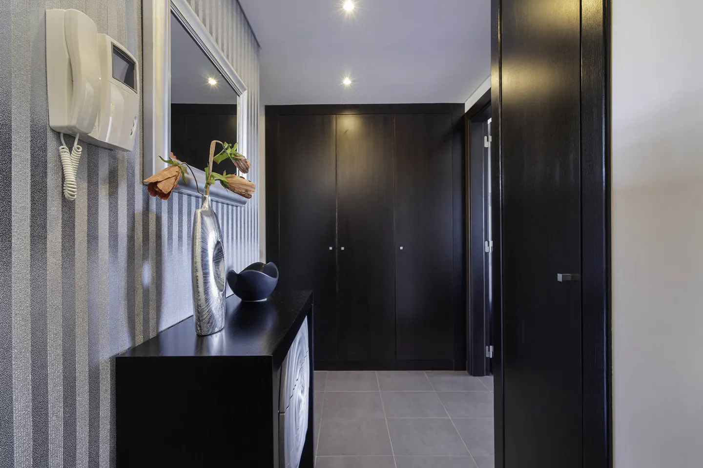 Hallway with striped wallpaper, black console table with vase and flowers, and dark wood closet doors.