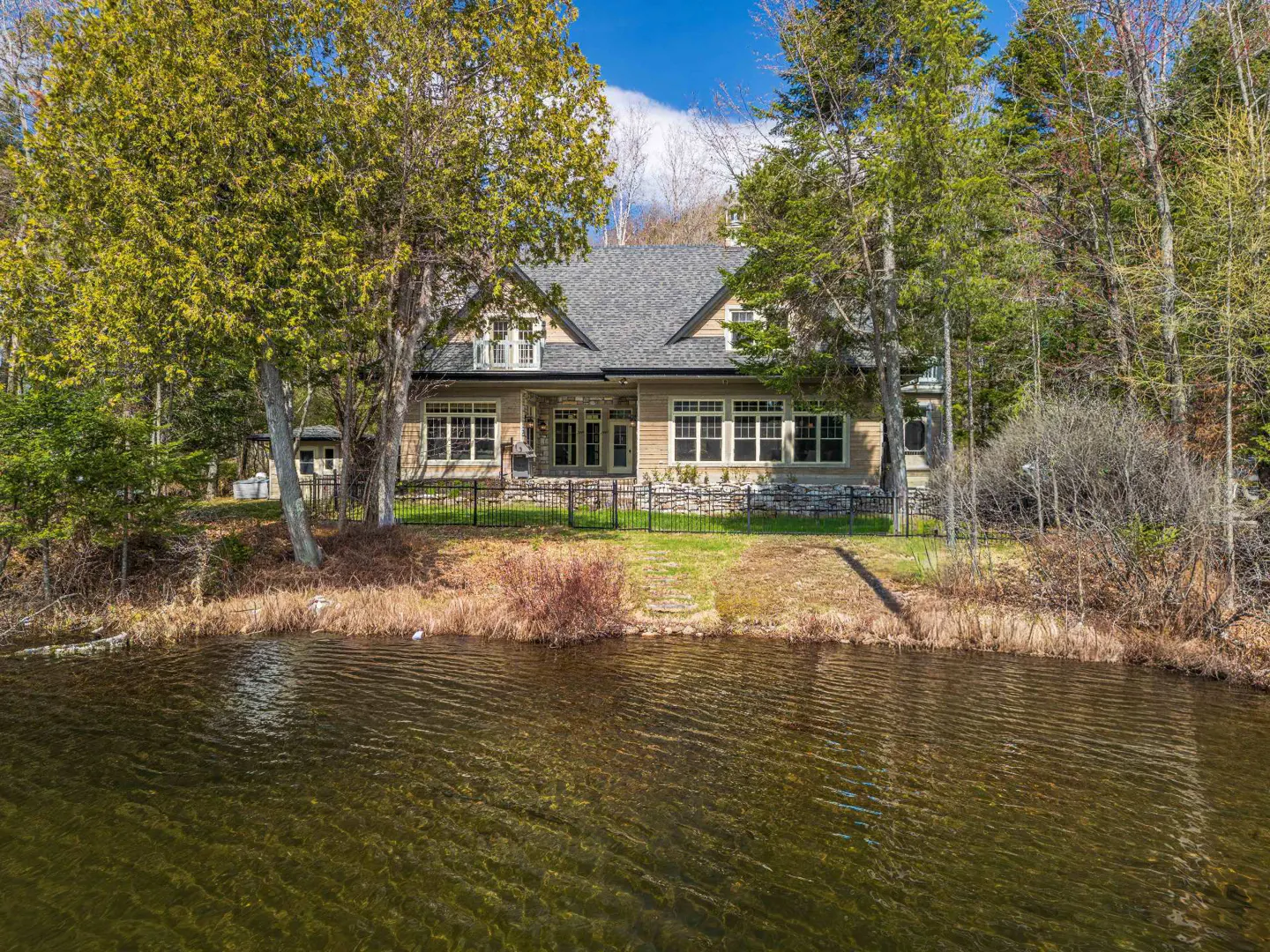 Lakefront home with a gray roof, tan siding, and black fence, surrounded by green trees and brown grass.