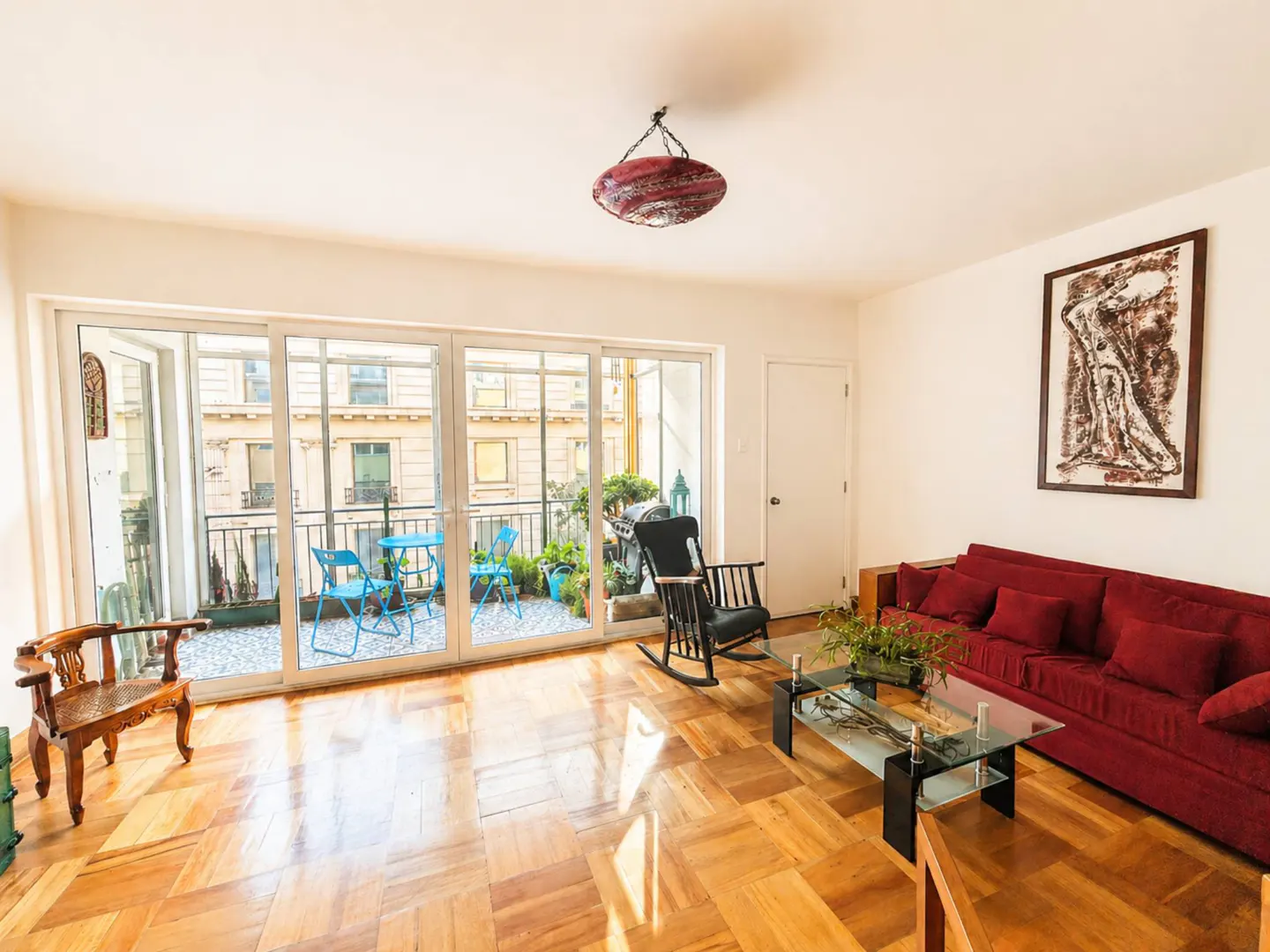 Bright living room with wood floors, red sofa, glass table, and balcony with blue furniture. Artwork on the wall.