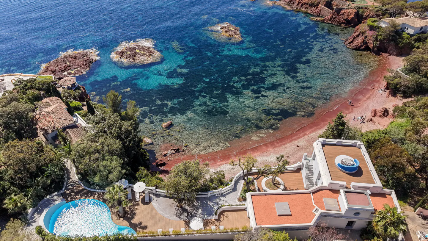 Aerial view of a luxury villa with a pool, overlooking a red sand beach and turquoise water.