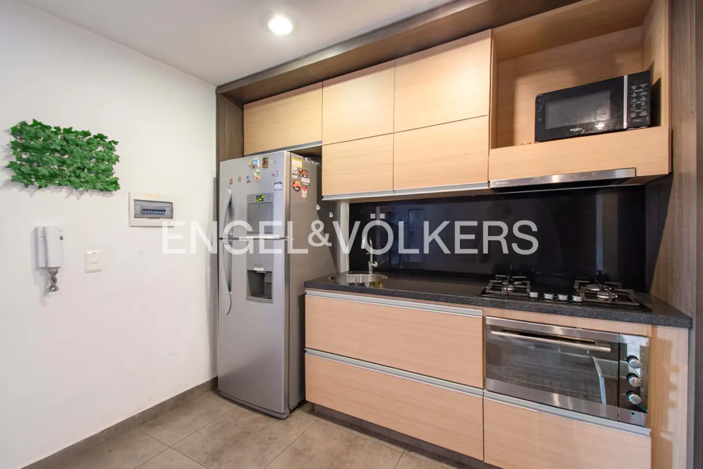 A modern kitchen with light wood cabinets, stainless steel appliances, and a black backsplash. A silver refrigerator stands next to the cabinets.