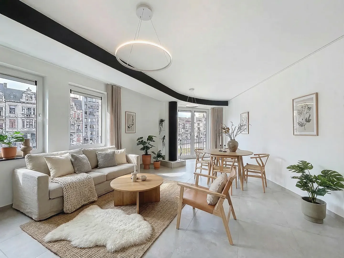 Bright, modern living room with a beige sofa, round wood table, and dining set. Plants and art add warmth to the white walls and gray floors.