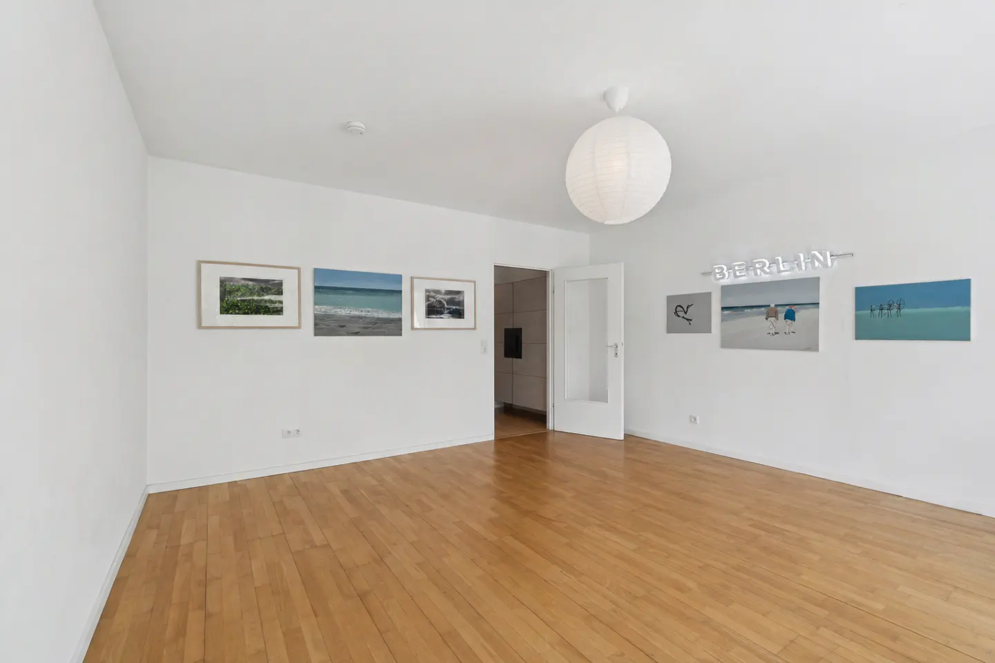 Bright, empty room with wood floors and white walls. Art hangs on the walls, including a neon "BERLIN" sign. A white paper lantern hangs from the ceiling.
