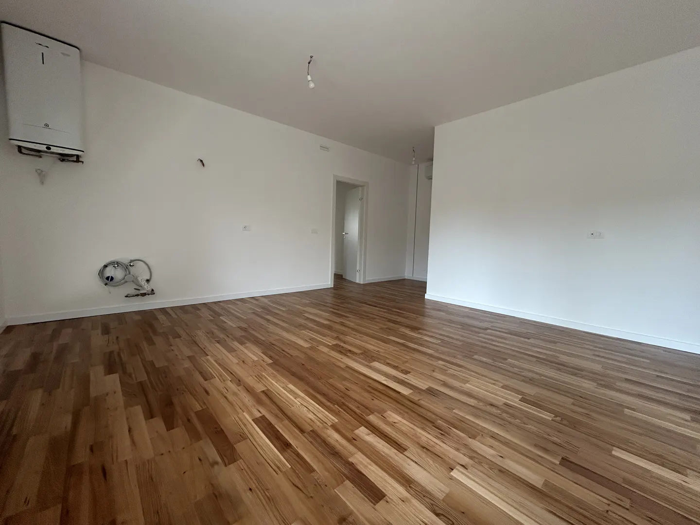 An empty room with light brown wood floors and white walls. A white water heater is mounted on the wall.