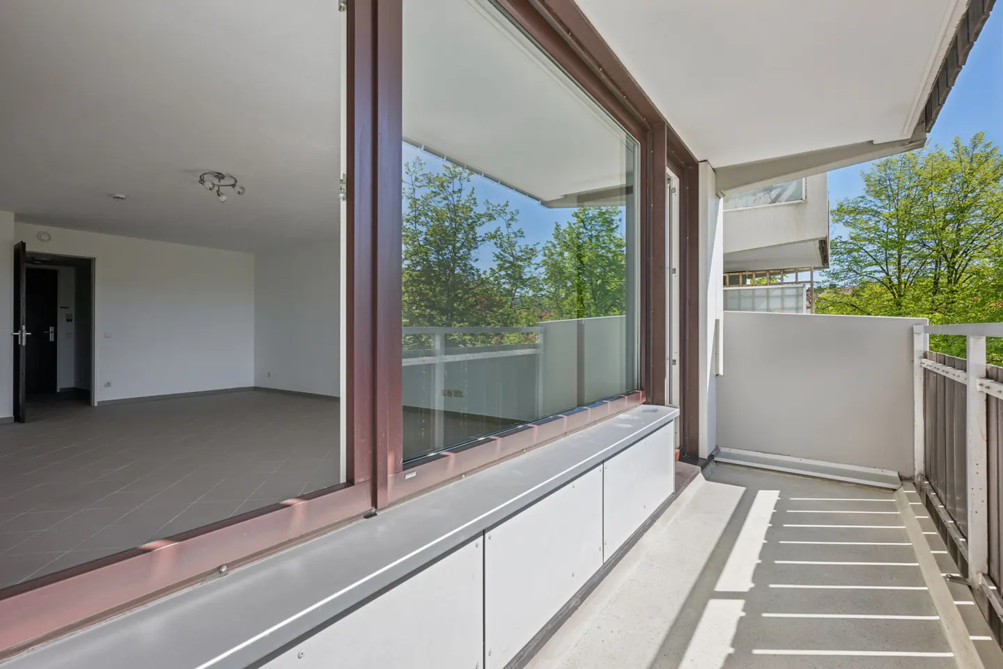 Balcony view with brown-framed windows looking into a bright, empty room. Outside, trees and a blue sky are visible. The balcony has a gray floor and metal railings.