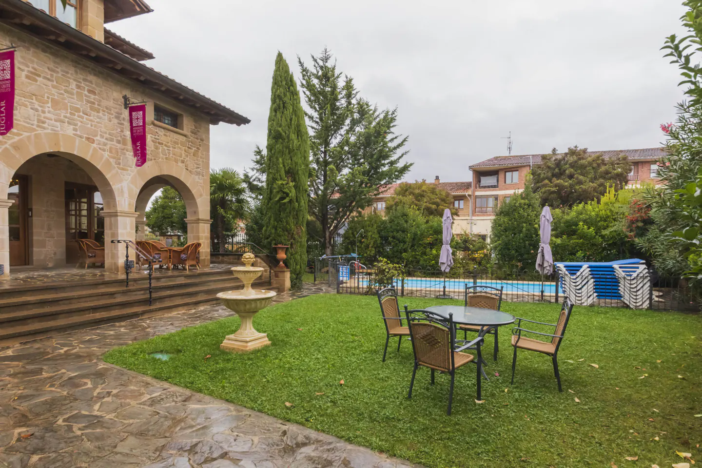 Stone building with arched porch, fountain, and lawn with table and chairs. A pool and stacked chairs are in the background.