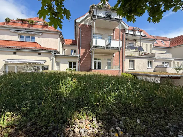Exterior view of a multi-story apartment building with balconies, red tile roofs, and green foliage in the foreground.