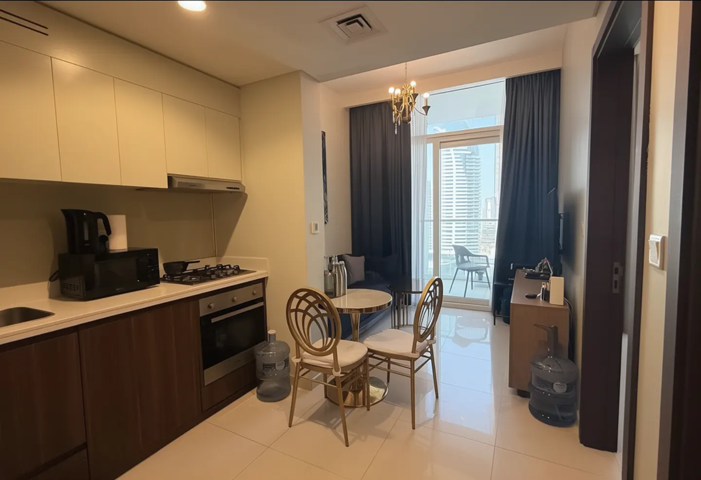 A modern apartment interior with a kitchen, dining area, and balcony with city views. The kitchen features light cabinets and stainless steel appliances.
