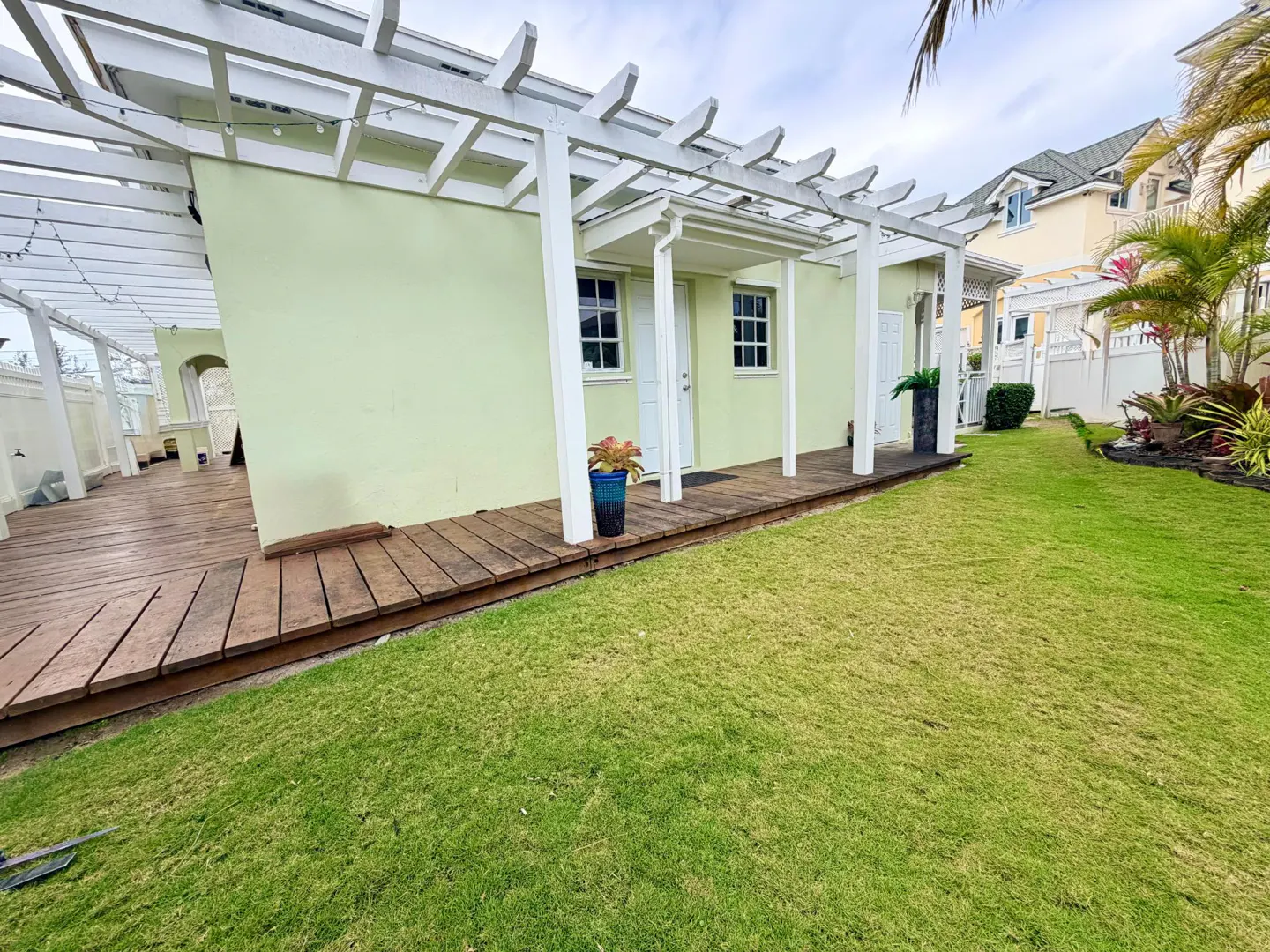 Exterior view of a light green house with a white pergola, wooden deck, and green lawn.