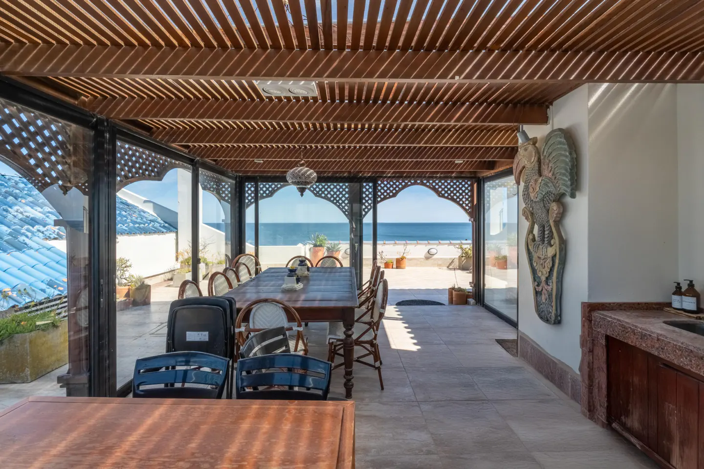 Indoor dining area with a long wooden table and chairs, glass walls, and a view of the ocean. A wooden pergola covers the ceiling.