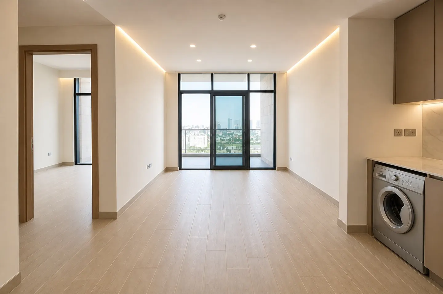 Bright, empty apartment with wood floors, a balcony with city views, and a silver washing machine next to kitchen cabinets.