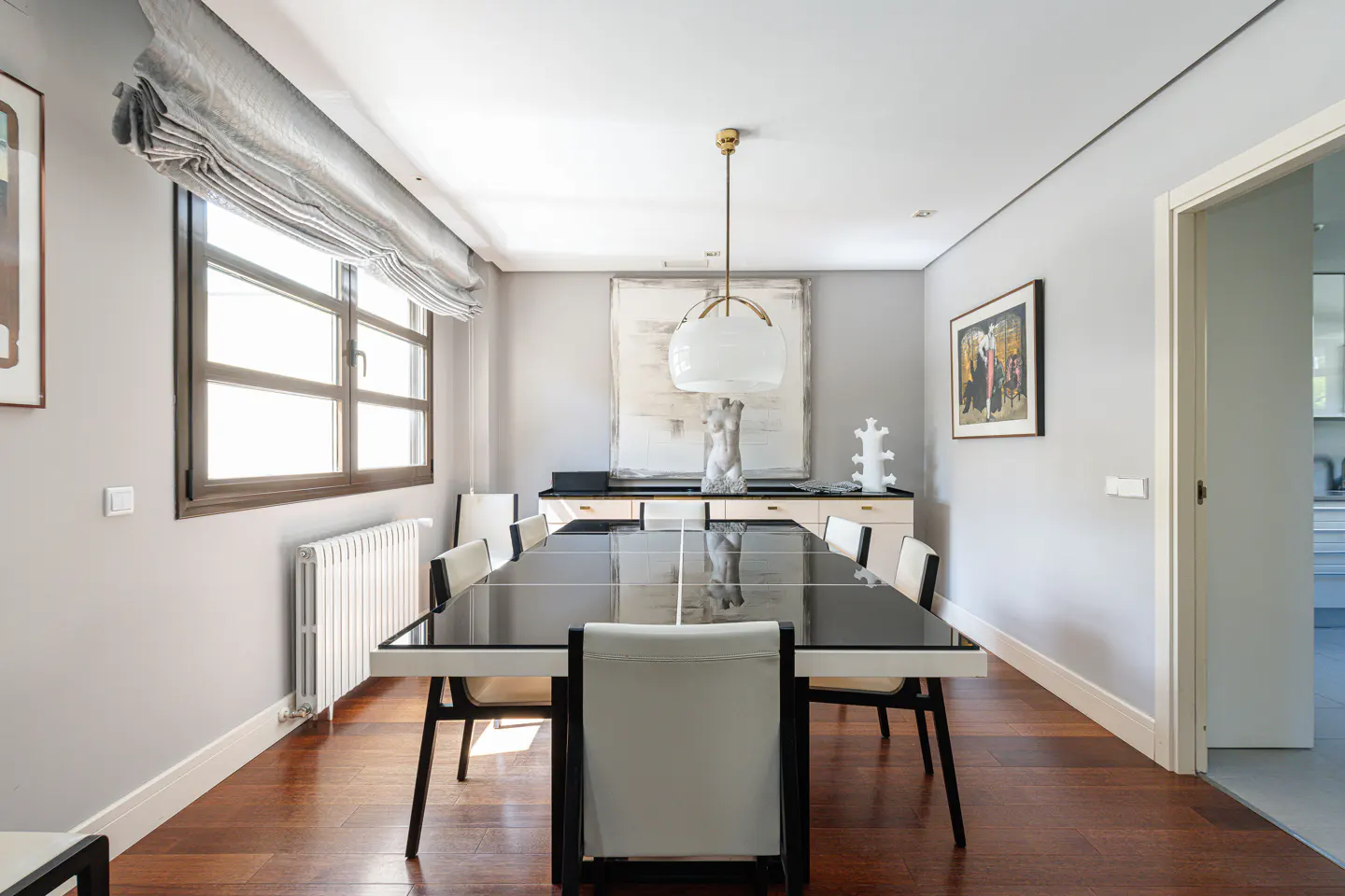 A modern dining room with a black table, white chairs, and hardwood floors. A window with gray blinds is on the left.
