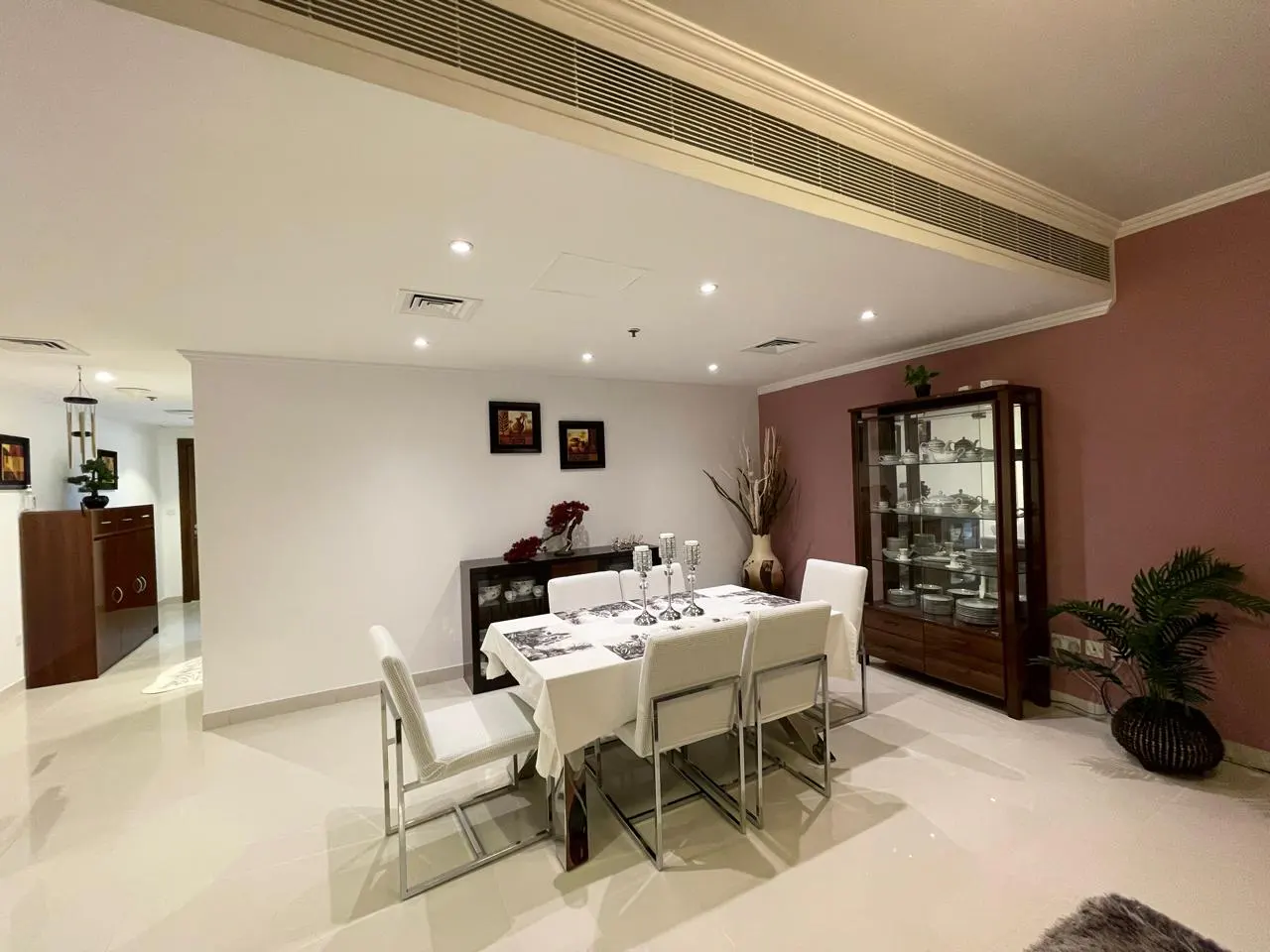 Dining room with white table and chairs, dark wood cabinet, and pink accent wall. The room is well-lit with recessed lighting.