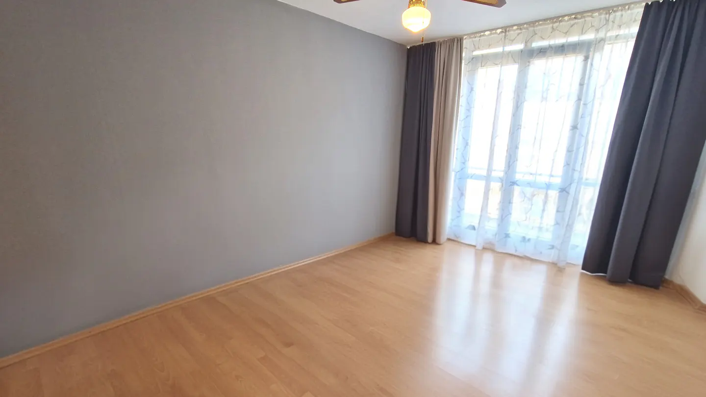 Empty room with gray wall, light wood floor, and window with sheer and dark gray curtains.