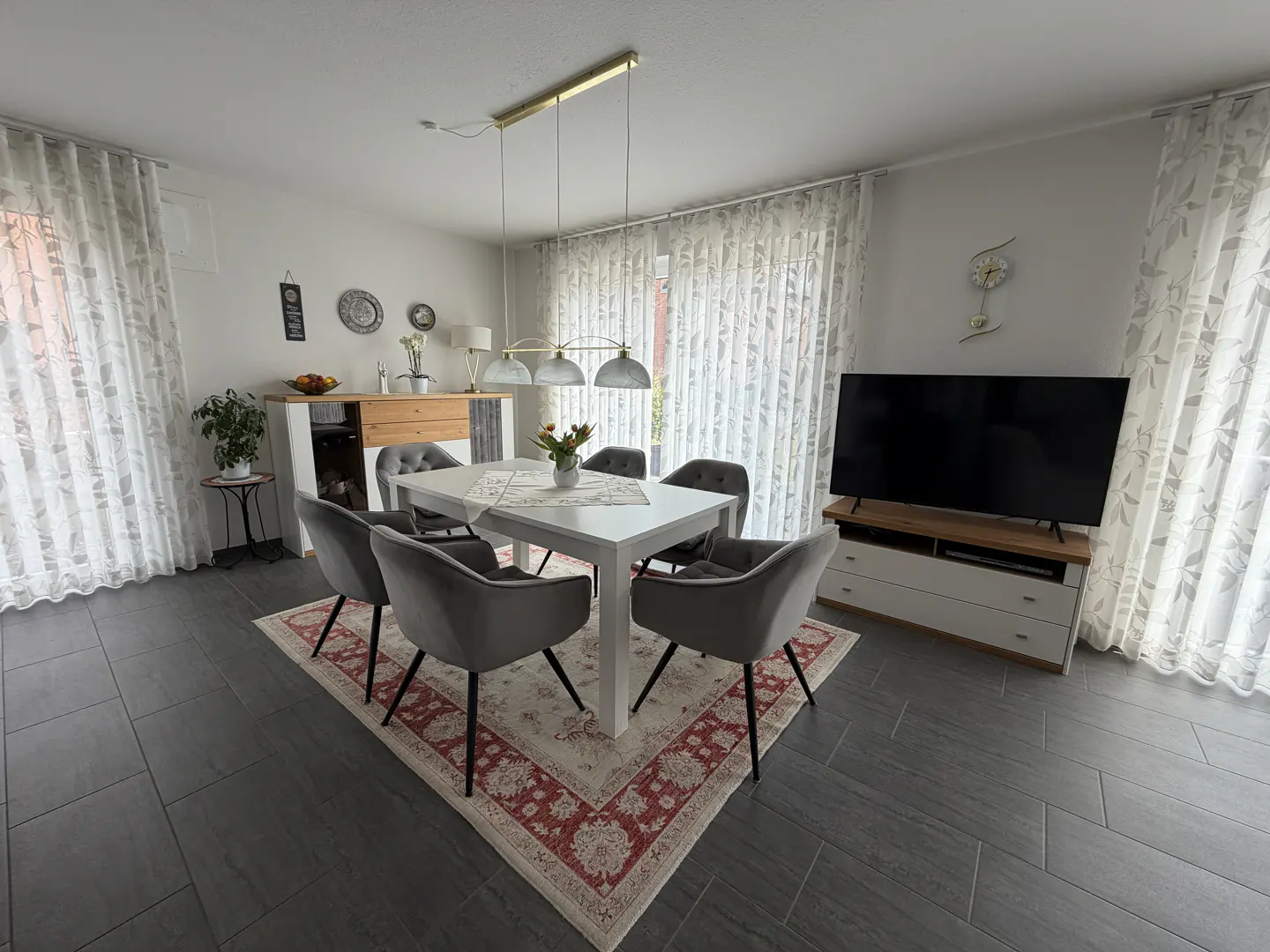 A dining room with a white table, six gray chairs, and a patterned rug. A TV and cabinet are against the wall.