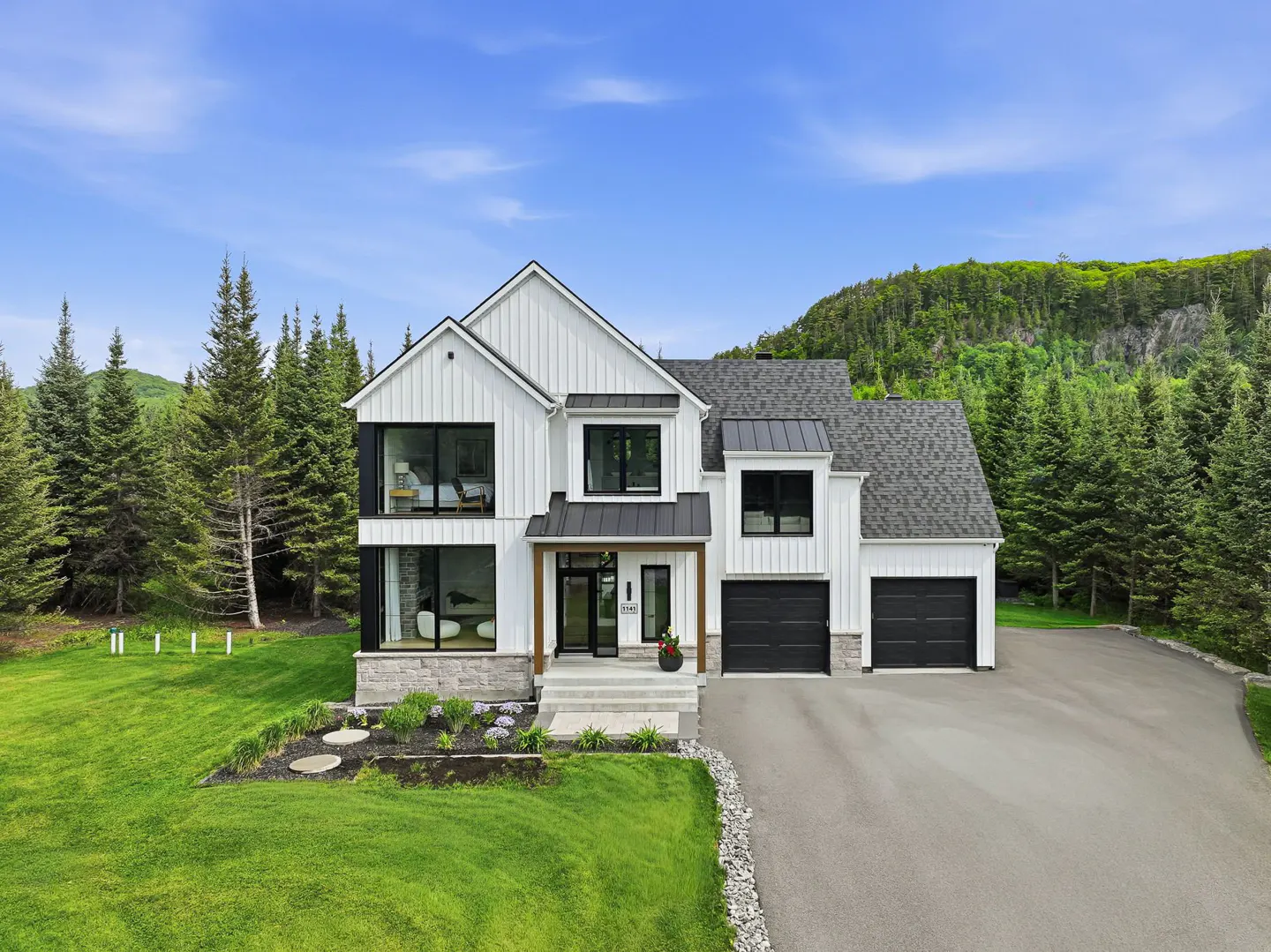 Modern white farmhouse with black accents, two-car garage, and stone base, set against a lush forest and blue sky.