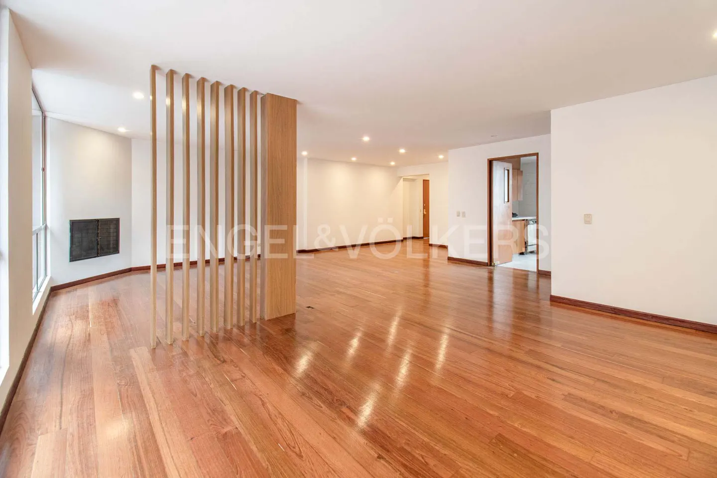 Bright, empty living room with hardwood floors, white walls, and a wooden slat divider. A fireplace is visible on the left.