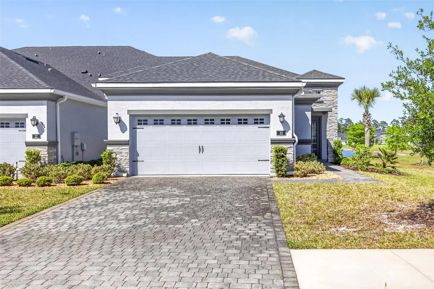 Exterior of a one-story home with a gray roof, white stucco, and a two-car garage. A brick driveway leads to the garage.