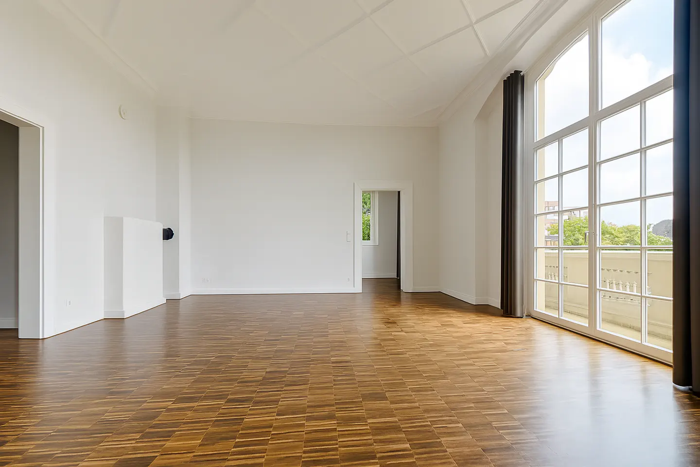 Empty room with parquet floor, white walls, and large windows with balcony view.
