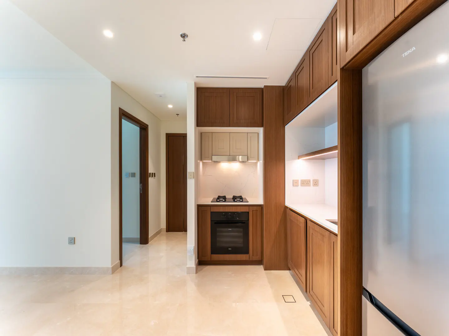 A bright, modern kitchen with wood cabinets, white countertops, and stainless steel appliances. A hallway leads to other rooms.