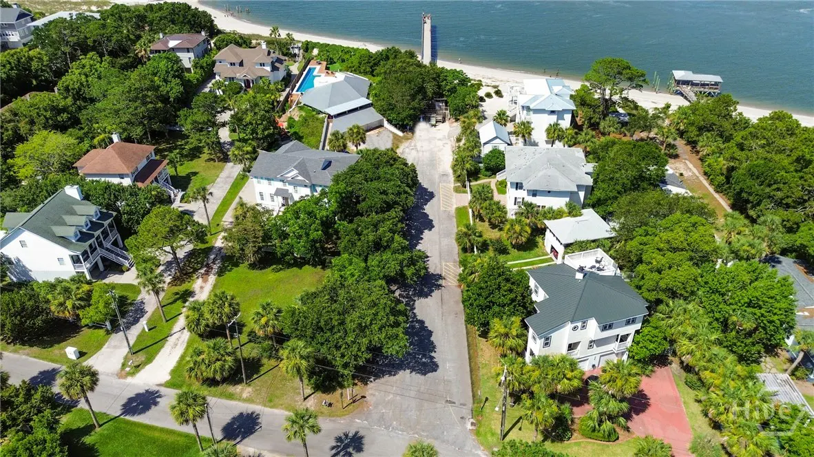 Aerial view of a coastal neighborhood with houses nestled among green trees, leading to a sandy beach and blue ocean.