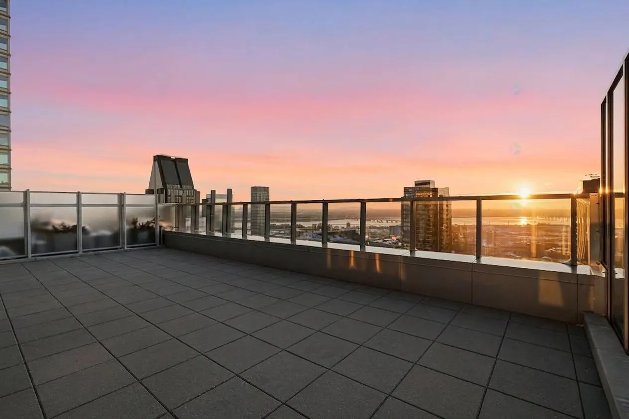 Rooftop patio with gray tiles and glass railings overlooking a city skyline at sunset with pink and blue sky.