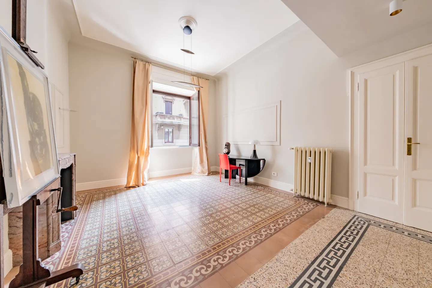 Bright room with patterned tile floor, easel with painting, desk with red chair, and window with peach curtains.