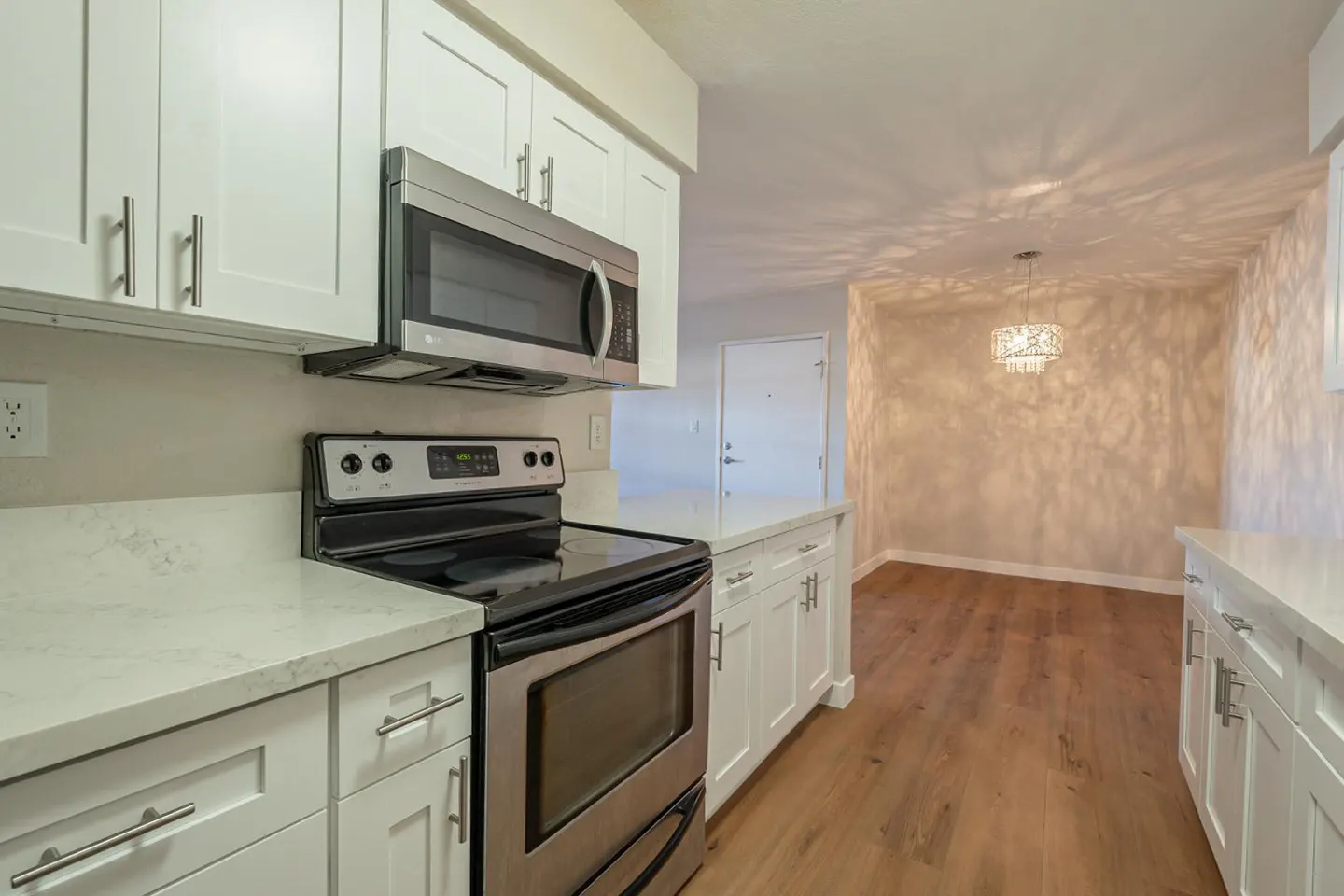 A bright kitchen with white cabinets, stainless steel appliances, and wood floors leading to a dining area with a chandelier.