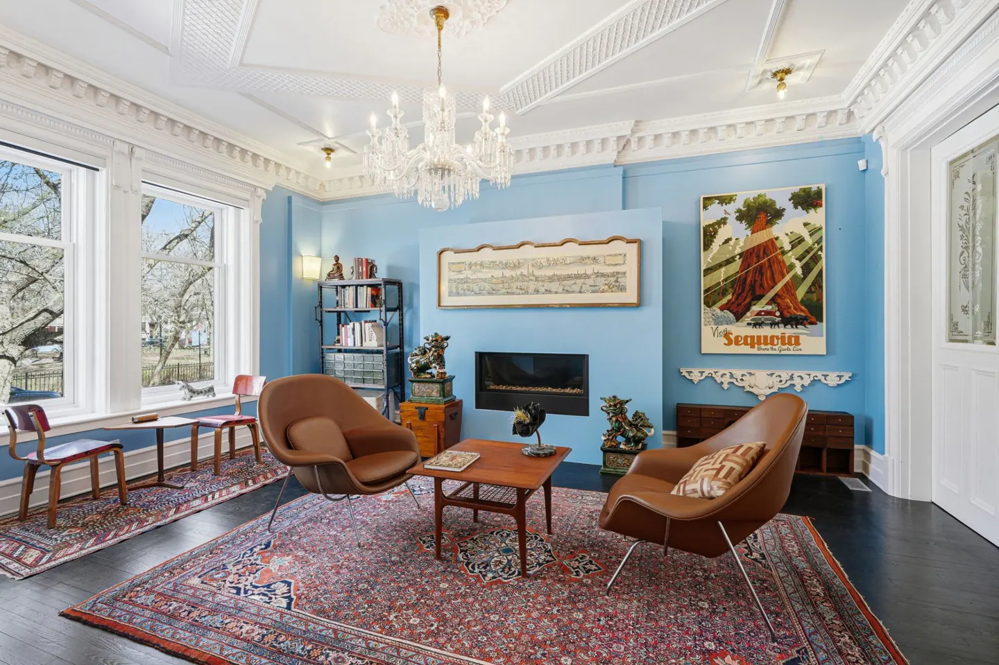 Living room with light blue walls, ornate white ceiling, chandelier, and two brown chairs on a red patterned rug. A fireplace and artwork are on the wall.