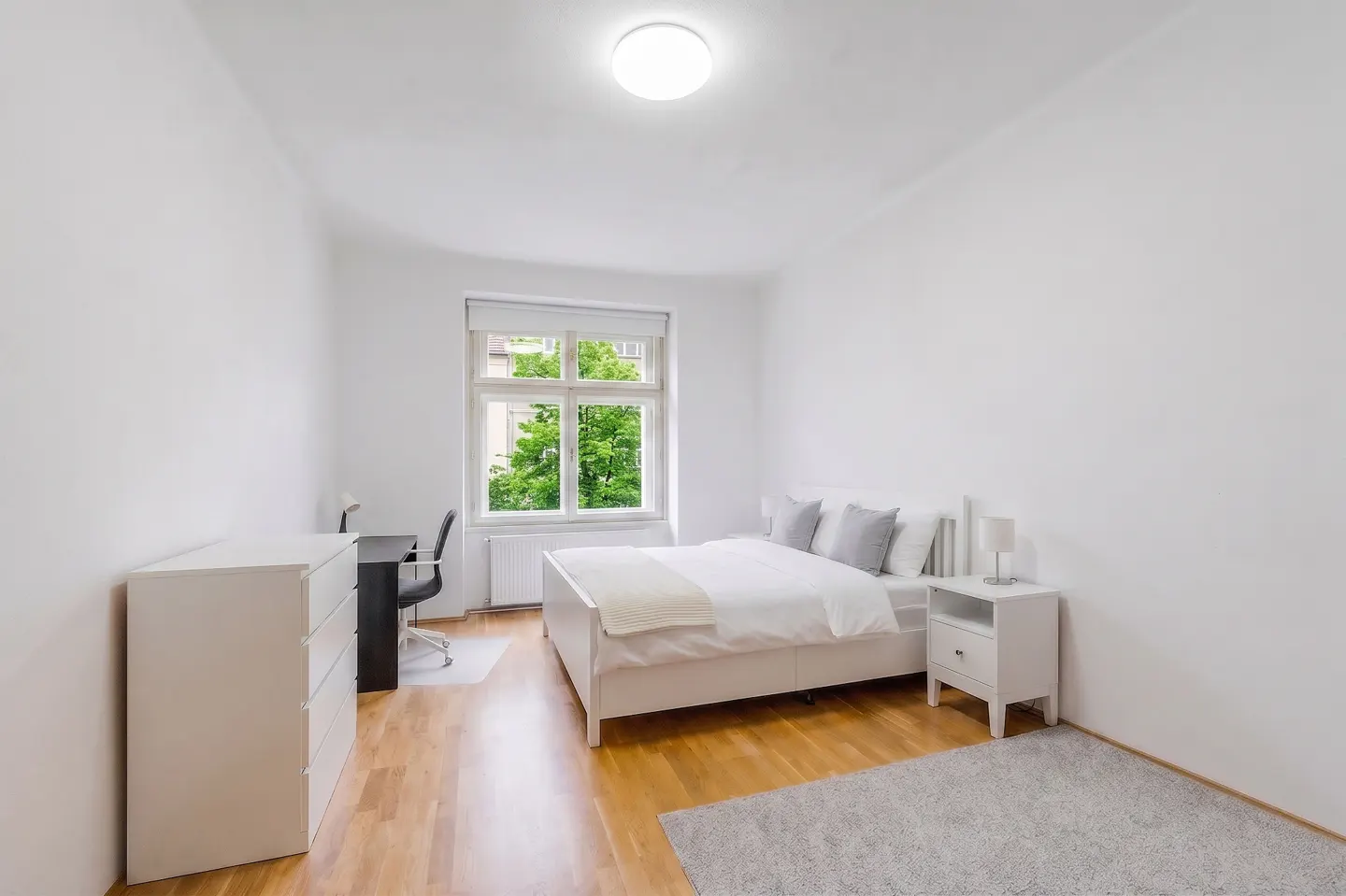 Bright bedroom with white walls, wood floor, and a large window. A white bed, desk, dresser, and gray rug furnish the space.