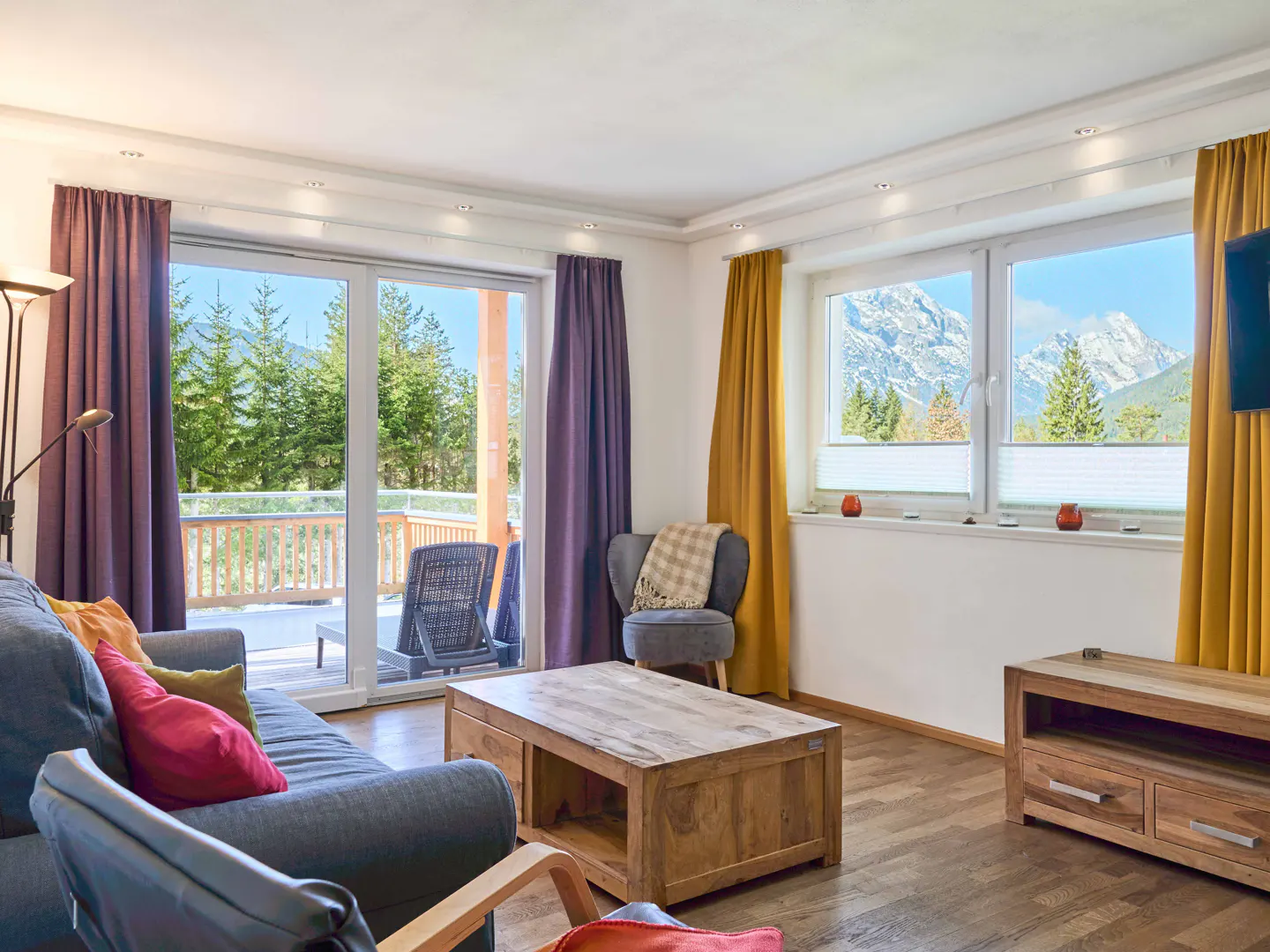 Living room with blue sofa, wooden table, and mountain view. Purple and gold curtains frame the windows and sliding glass door.
