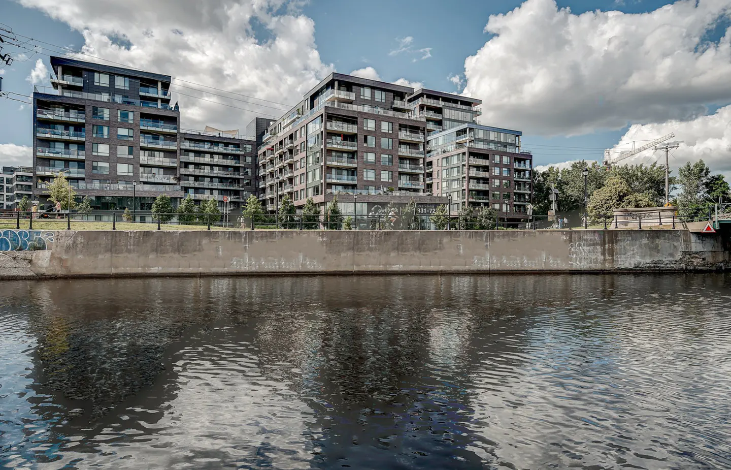 Modern brick apartment buildings with balconies line a canal under a cloudy sky. Water reflects the buildings and sky.