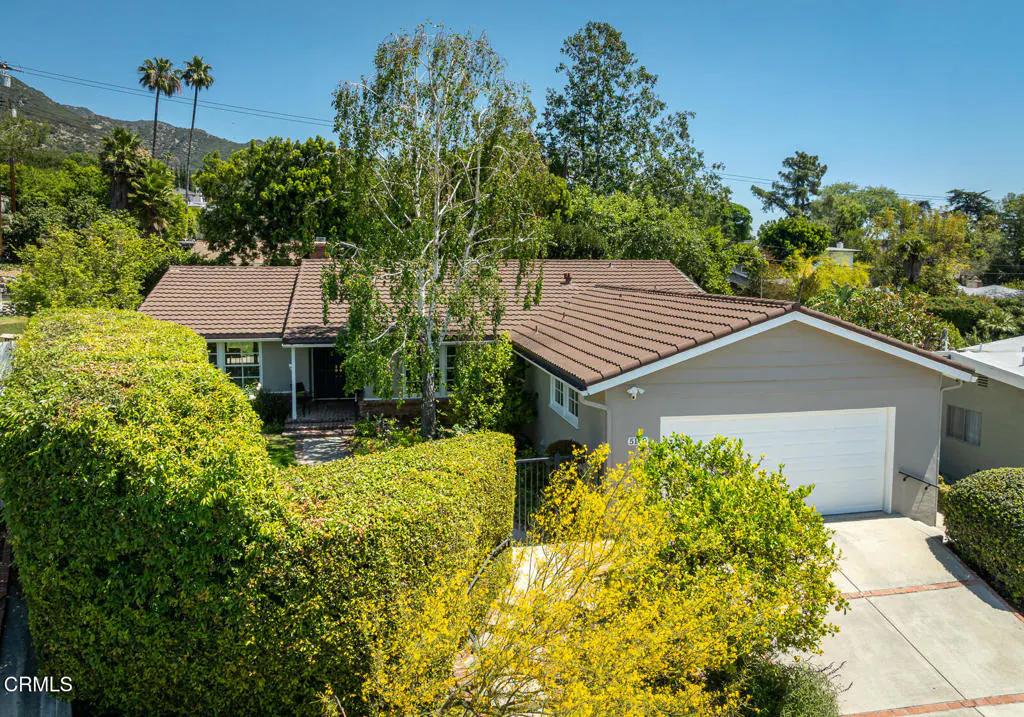 An eye-level shot of a single-story house with a brown roof, beige siding, and a white garage door, surrounded by lush green trees.