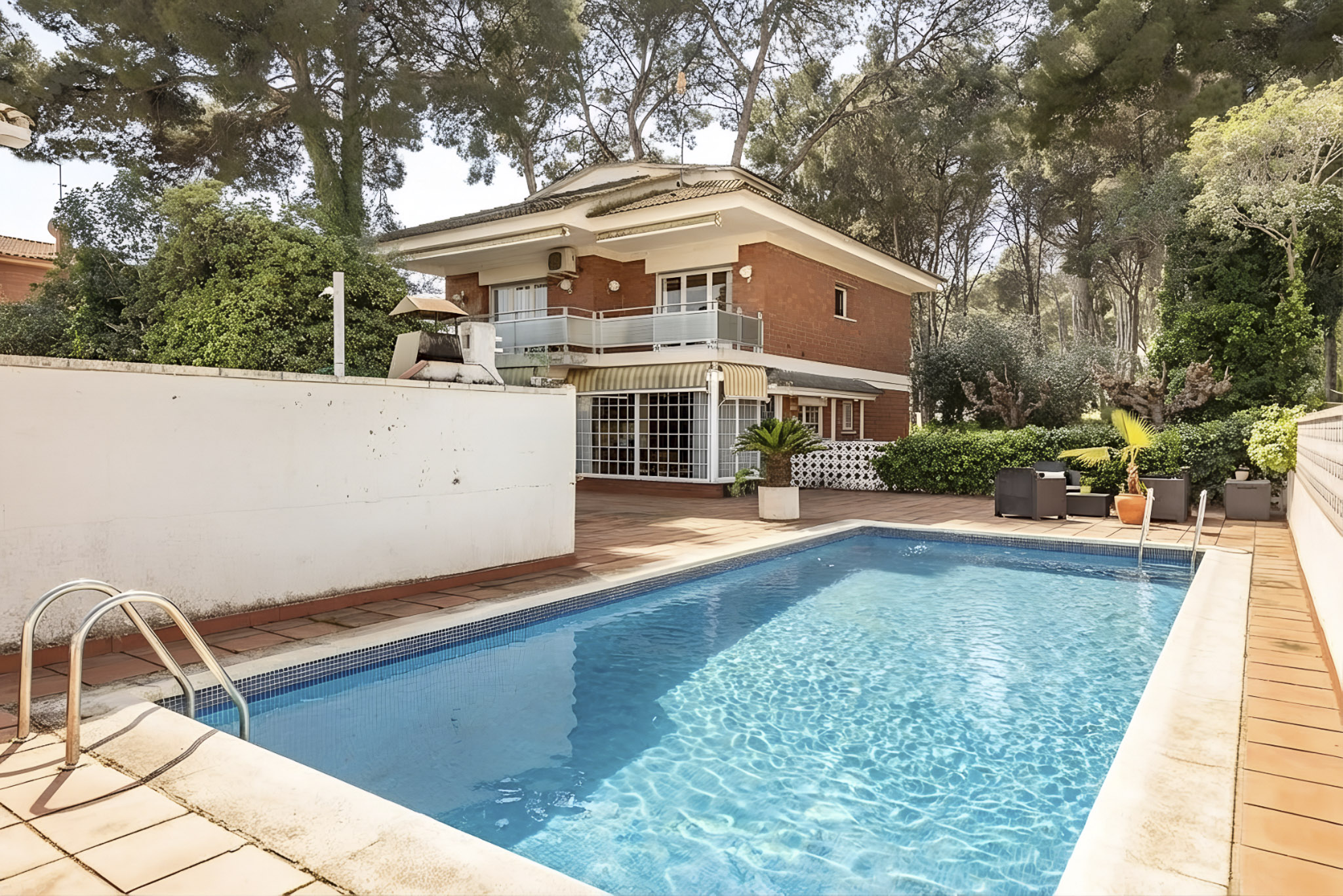 A two-story brick house with a blue tiled pool in the foreground. Patio furniture sits near the pool, surrounded by trees and greenery.