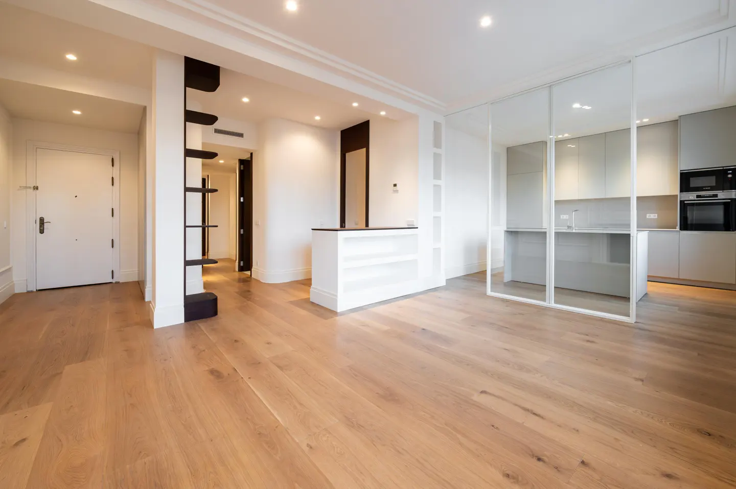 Bright, modern apartment with light wood floors. White walls, black shelving, and a kitchen visible through a glass partition.