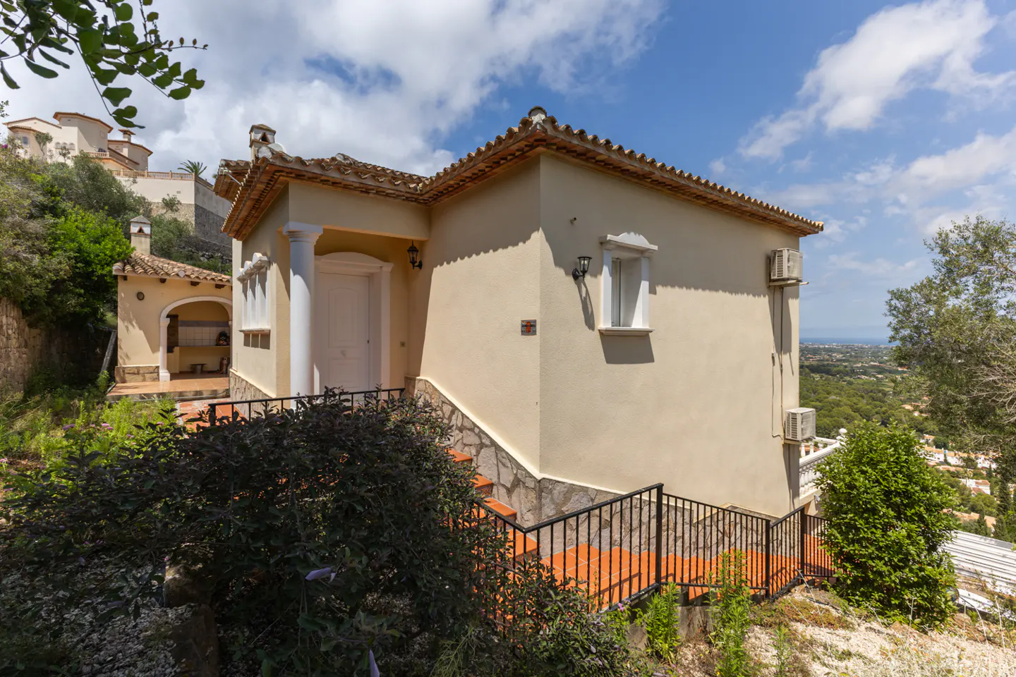 Cream-colored Mediterranean villa with a tiled roof, white columns, and terracotta stairs, set in a sunny hillside garden.