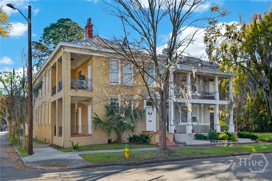 Two-story brick houses with balconies, one yellow and one gray, stand on a tree-lined street under a blue sky.