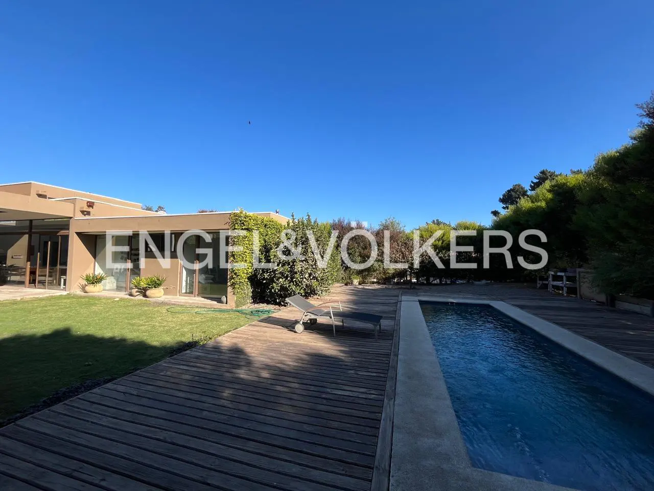 Exterior view of a modern tan house with a pool and wooden deck under a clear blue sky. A lounge chair sits on the deck.