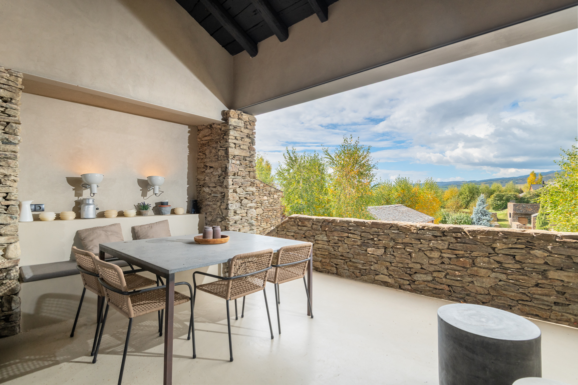 Outdoor patio with stone pillars, table, and chairs overlooking a scenic landscape with trees and cloudy sky.