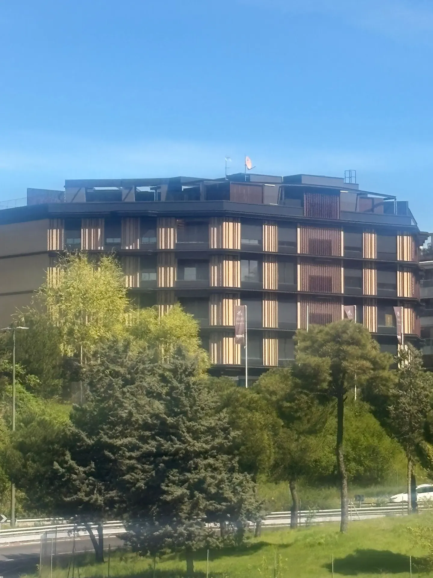 Modern apartment building with brown vertical slats, set against a blue sky and green trees.